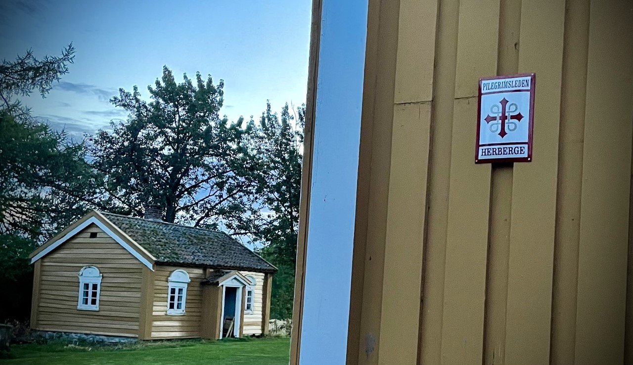 Lite gult trehus sett fra døra ved et skilt for pilegrimsherberge