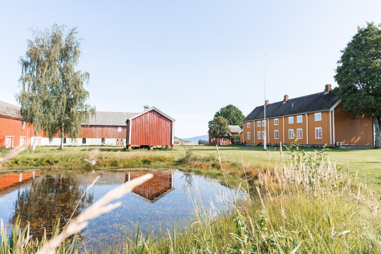 Stadsbygd prestegård med gule hovedbygninger og røde uthus speilet i en dam